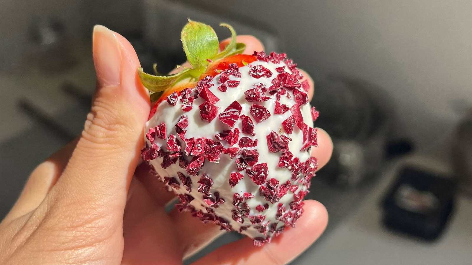 Mão segurando um morango cravejado, coberto por chocolate branco e pedaços de calda vermelha crocante